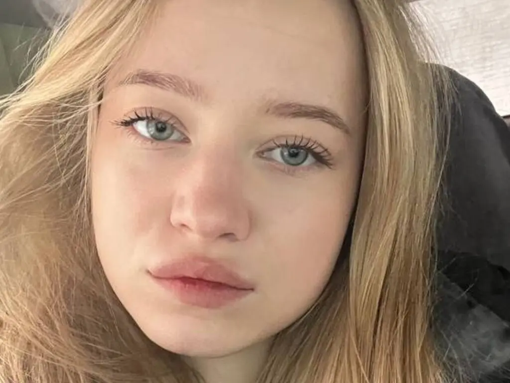 Close-up portrait of a blonde woman with blue eyes looking calmly at the camera in soft natural lighting.