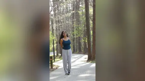 Smiling woman in a navy tank top and grey sweatpants walking on a sunny forest path.