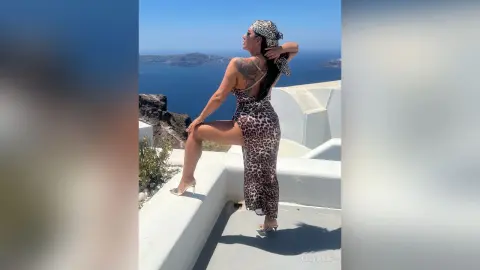 MissyJolie poses on a white balcony overlooking the ocean wearing a tight leopard print dress and white heels.