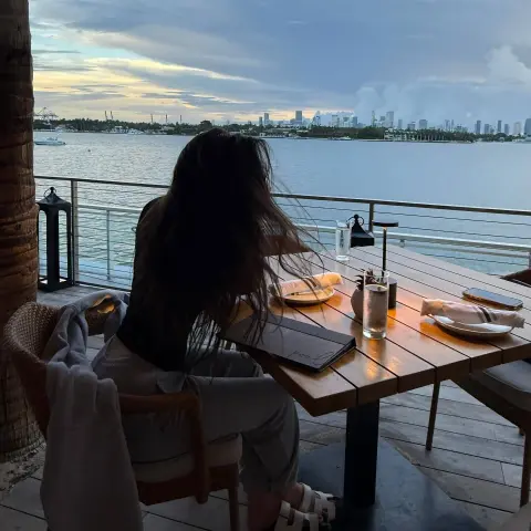 Woman in casual attire sitting at an outdoor waterfront restaurant table during a moody twilight sunset.