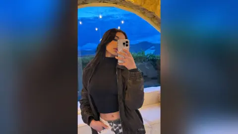 Young woman in a black turtleneck and leather jacket taking a selfie in a scenic outdoor setting at dusk.