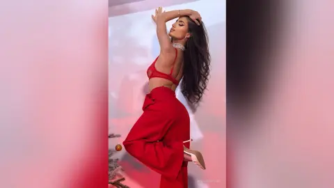 Elegant woman in red silk trousers and lace bralette posing confidently against a soft pink background.