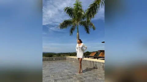 Aleja stands on a stone patio wearing a white long-sleeved mini dress, posing next to a tall palm tree under a blue sky.