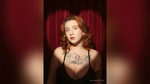 Redheaded woman in a black dress posing under a heart-shaped light against a red velvet curtain backdrop.