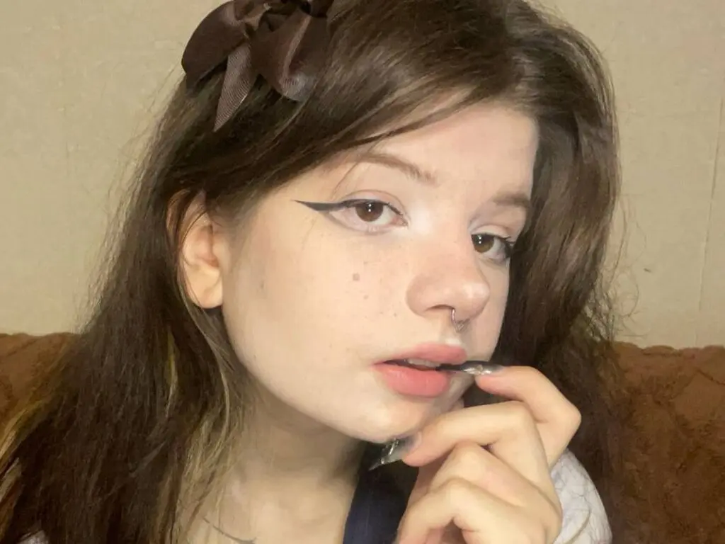 Young woman with winged eyeliner and a brown hair bow posing in a cozy indoor setting.