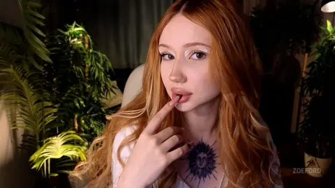 Redheaded woman in a white top with a sun tattoo posing thoughtfully near indoor green plants.