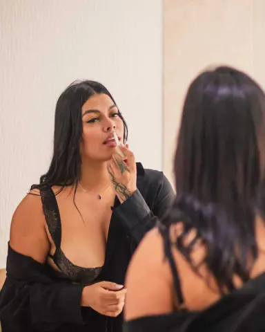 Young woman with long dark hair applying lip gloss in a mirror while wearing a black lace camisole.