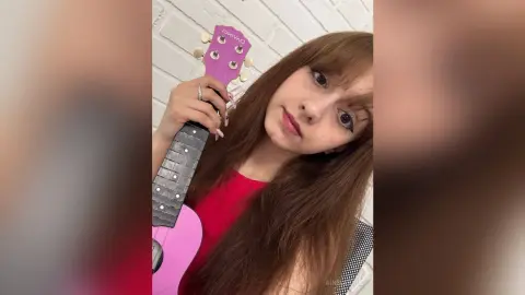 Young woman with long brown hair holding a pink acoustic guitar against a white brick wall.