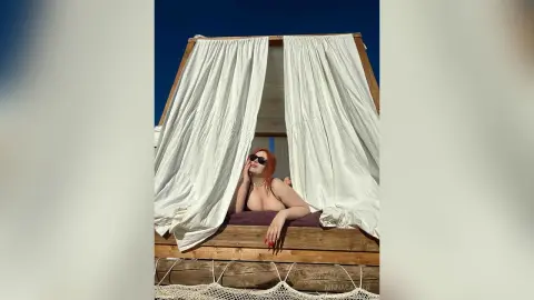 Redheaded woman in sunglasses relaxing in a white draped cabana under a clear blue sky.