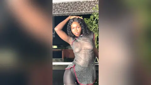 Confident Black woman in a sheer silver mesh top posing on a balcony at night.
