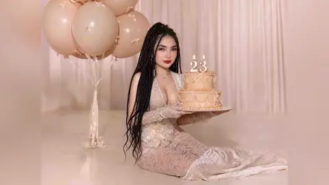 Young woman with long braids in a lace gown celebrating her 23rd birthday with a cake and balloons.