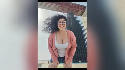 Smiling woman with curly hair in a pink cardigan and white tank top standing outdoors by a modern skyscraper.