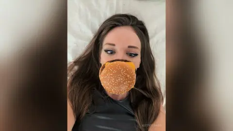 Young woman with long brown hair holding a large hamburger in a casual indoor setting.