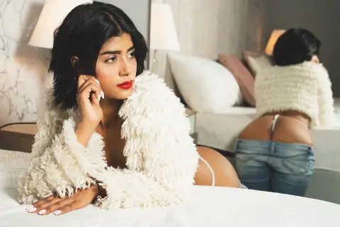 Glamorous woman in a white faux fur coat lying on a bed in a cozy, moody bedroom setting.