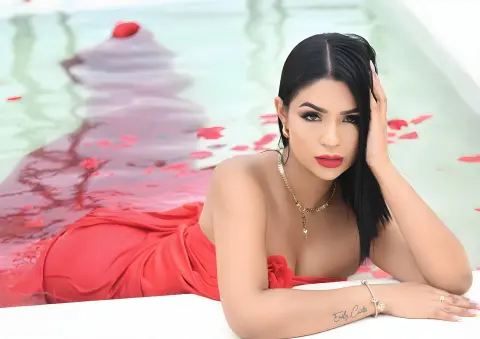 Emilycurtiis with long black hair wearing a red dress while relaxing in a pool filled with red rose petals.