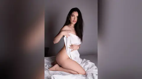 IrisOwen poses naked on white sheets, holding a white sheet against her chest and exposing her bare breasts and stomach.
