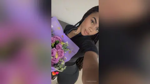 PamelaCreed poses with a bouquet of purple roses, wearing a black ribbed top and showing her long dark braids.