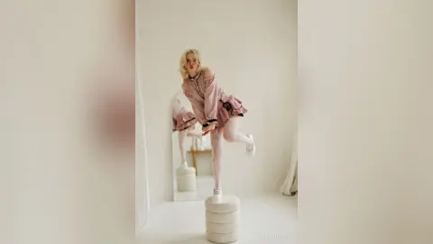 LynGoodhart poses in a pink ruffled dress and white platform shoes while standing on a white pedestal in a bright room.