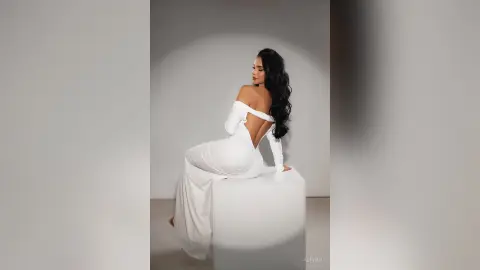 Alison poses sitting on a white block wearing a long, white off-the-shoulder dress that shows her bare back.