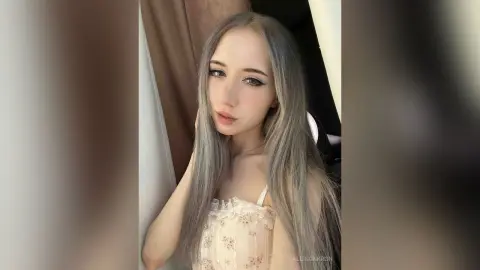 Young woman with long silver hair wearing a white lace corset top in a soft, dreamy indoor setting.