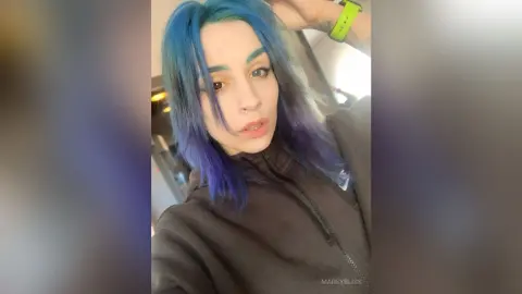 Young woman with blue hair wearing a black zip-up hoodie in a casual indoor setting.