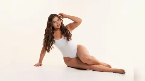 Curvy woman in a white bodysuit posing gracefully on a white background in a minimalist studio setting.