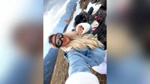 Anita poses outdoors in a denim jacket and white furry headband, showing her long blonde hair against a mountain landscape.