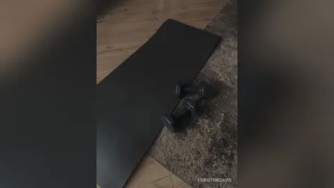 ChristineDavis poses on a black yoga mat with two black dumbbells on a grey rug over a wooden floor.