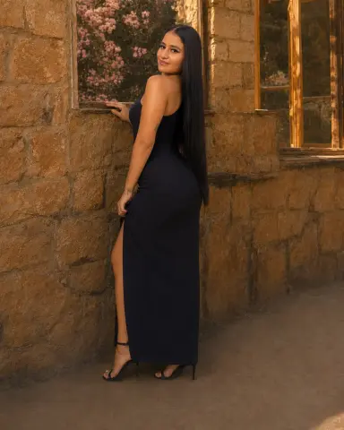 Elegant woman in a long black evening dress posing by a rustic stone wall in warm sunlight.