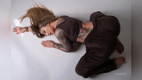 Tattooed woman in brown loungewear lying on a white floor in a relaxed, moody studio setting.