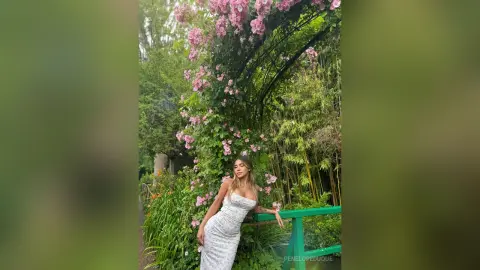 Elegant woman in a white floral dress posing in a lush green garden with pink blooming flowers.