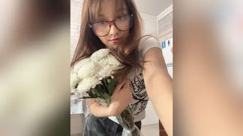 Young woman with glasses holding a white flower bouquet in a bright indoor setting with a soft, dreamy mood.