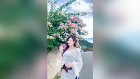 EnyaLin stands outdoors next to pink flowers wearing a white mesh long-sleeve top and a black belt.
