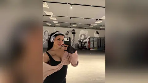 NicoleBella takes a mirror selfie wearing white headphones and a low-cut black top with pink sleeves.