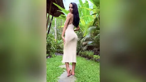 Elegant woman in a white slip dress standing on a garden path in a lush tropical jungle setting.