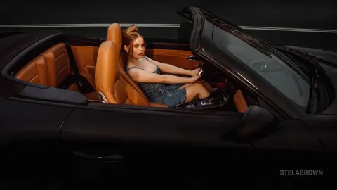 StelaBrown sits in the tan leather seat of a black convertible, wearing a denim top and looking toward the camera.