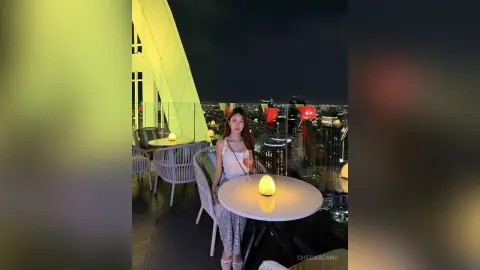 CheCascano sits at a rooftop table at night, wearing a white tank top and patterned pants while holding a drink.