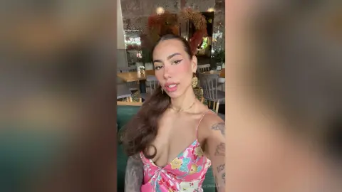 VictoriaFontaine poses in a pink floral top with her long brown hair swept to one side, showing off her tattoos and gold earrings in a restaurant setting.