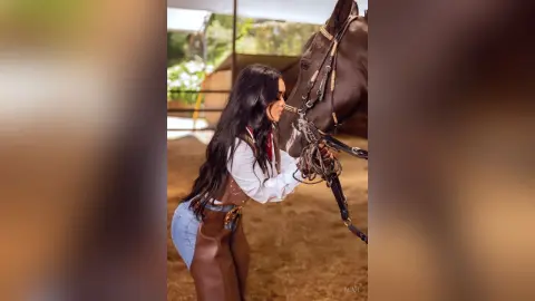 Woman in white blouse and denim shorts bonding with a brown horse in a rustic ranch setting.