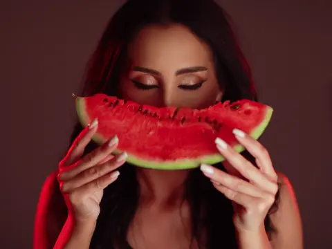 Young woman with long dark hair holding a fresh watermelon slice in front of her face against a red background.