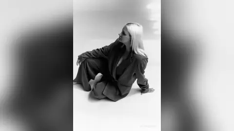 Blonde woman in a stylish oversized pinstripe suit sitting on a white floor in a moody black and white portrait.