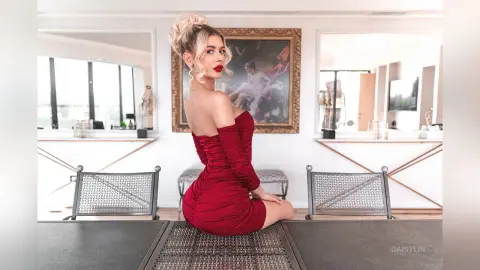 DaisyLin poses in a red off-the-shoulder dress, looking over her shoulder with blonde hair styled in a bun.