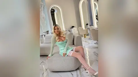 Blonde woman in a mint green long-sleeve mini dress relaxing on a grey sofa in a bright, modern luxury living room.