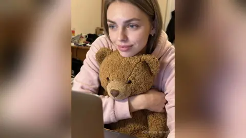 CharlotteReimer wearing a pink hoodie while hugging a brown teddy bear and looking at her laptop.