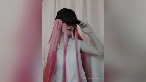 Woman with long pink hair wearing a white sleeveless top and black cap in a soft indoor setting.
