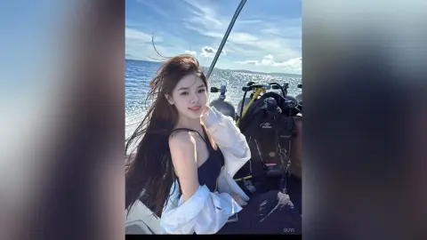 Smiling woman in a black swimsuit and white shirt relaxing on a boat under a sunny blue sky.