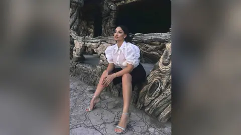 AnnaKarev sits on a stone ledge wearing a white puffy blouse, black skirt, and silver high heels.