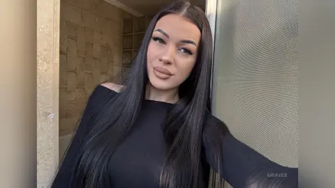 AriaVex poses with long straight black hair wearing a black long-sleeved top against a textured wall.