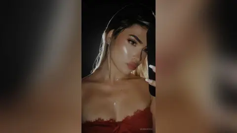 Glamorous woman in a red lace top taking a moody selfie with glowing makeup in dim lighting.