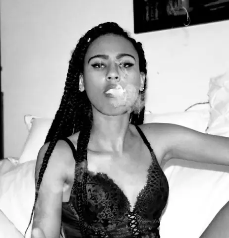 GoddessHoneyXO — GoddessHoneyXO with long braids wearing a lace bodysuit while blowing smoke in her bedroom.